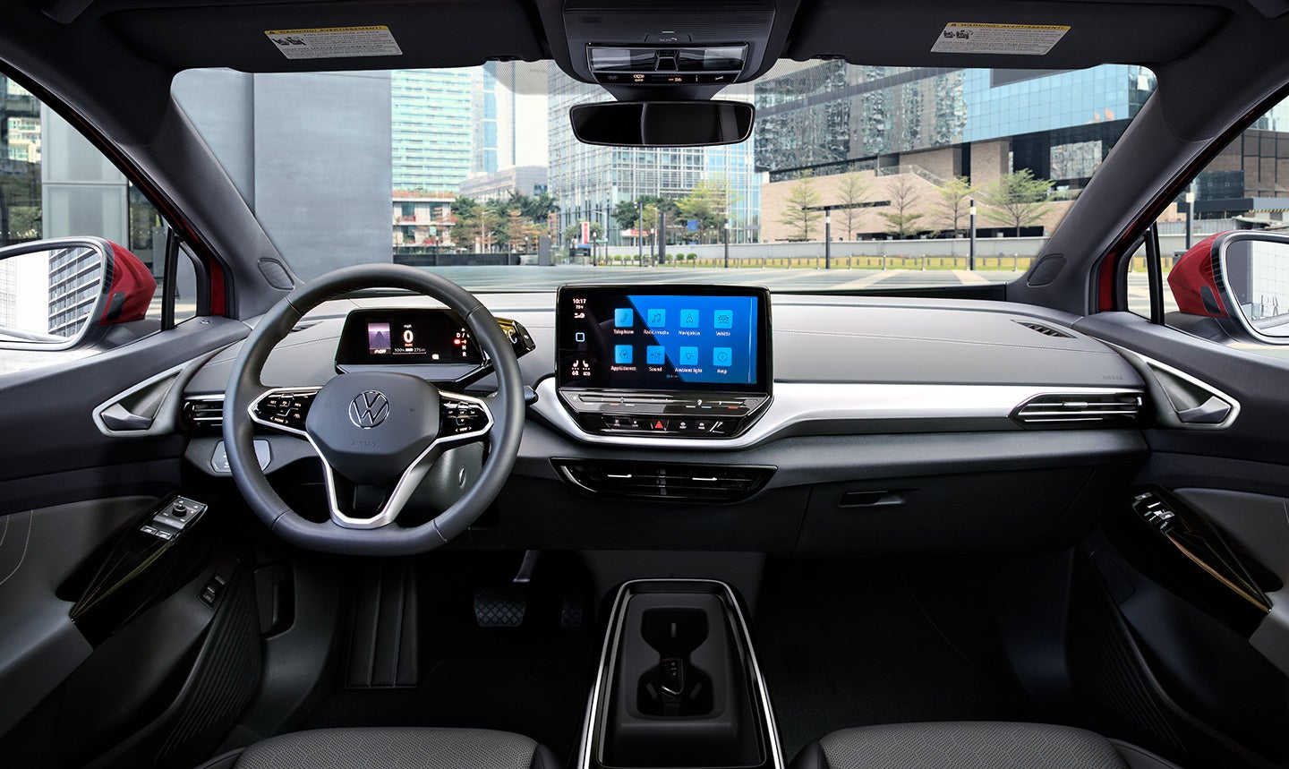 An interior shot of the cockpit area in an ID.4 AWD Pro S Plus shows the steering wheel, driver display, and center console. Parked downtown, a modern city can be seen through the windshield.